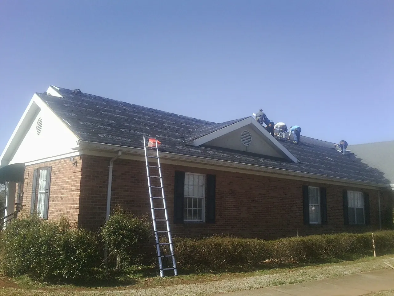 Crew tearing shingles off a brick ranch roof with a ladder set — Emergency Roof Repair work by our crew in Rigby