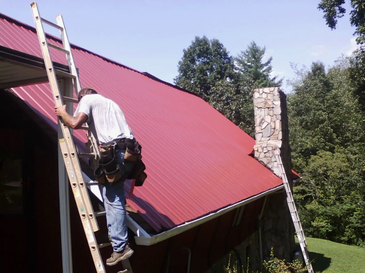 Roofer on a ladder beside a red metal roof and stone chimney — Soffit & Fascia Repair by Roofing Brothers in Rigby